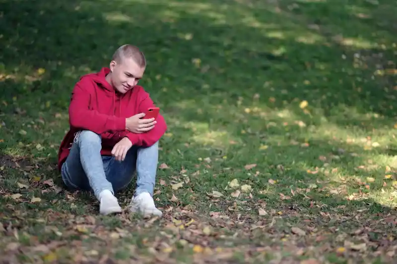 Ung kille sitter i gräset och tittar på mobiltelefon.